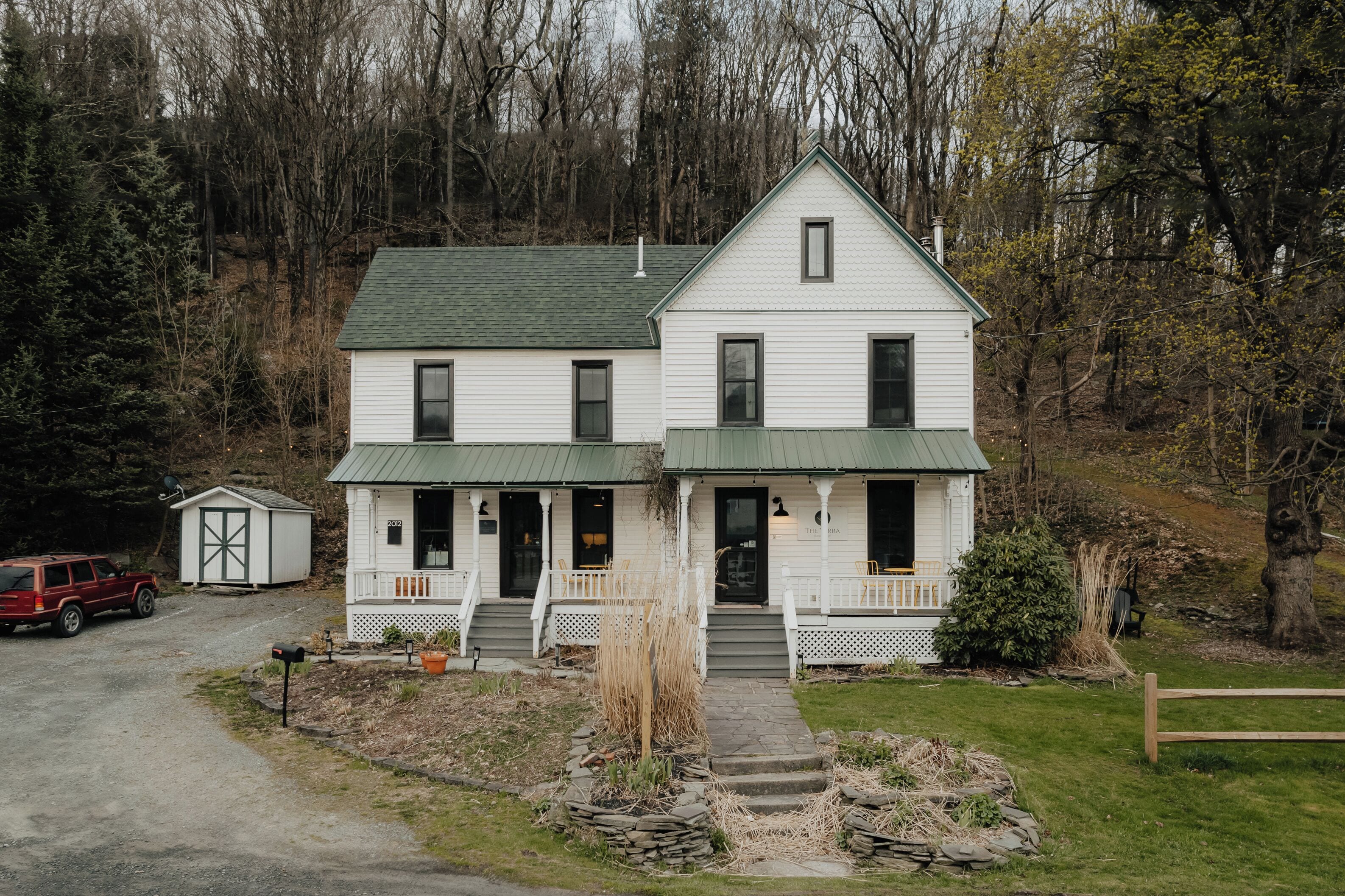 A two-story white house with a green roof, a porch, and a small shed in the front yard.