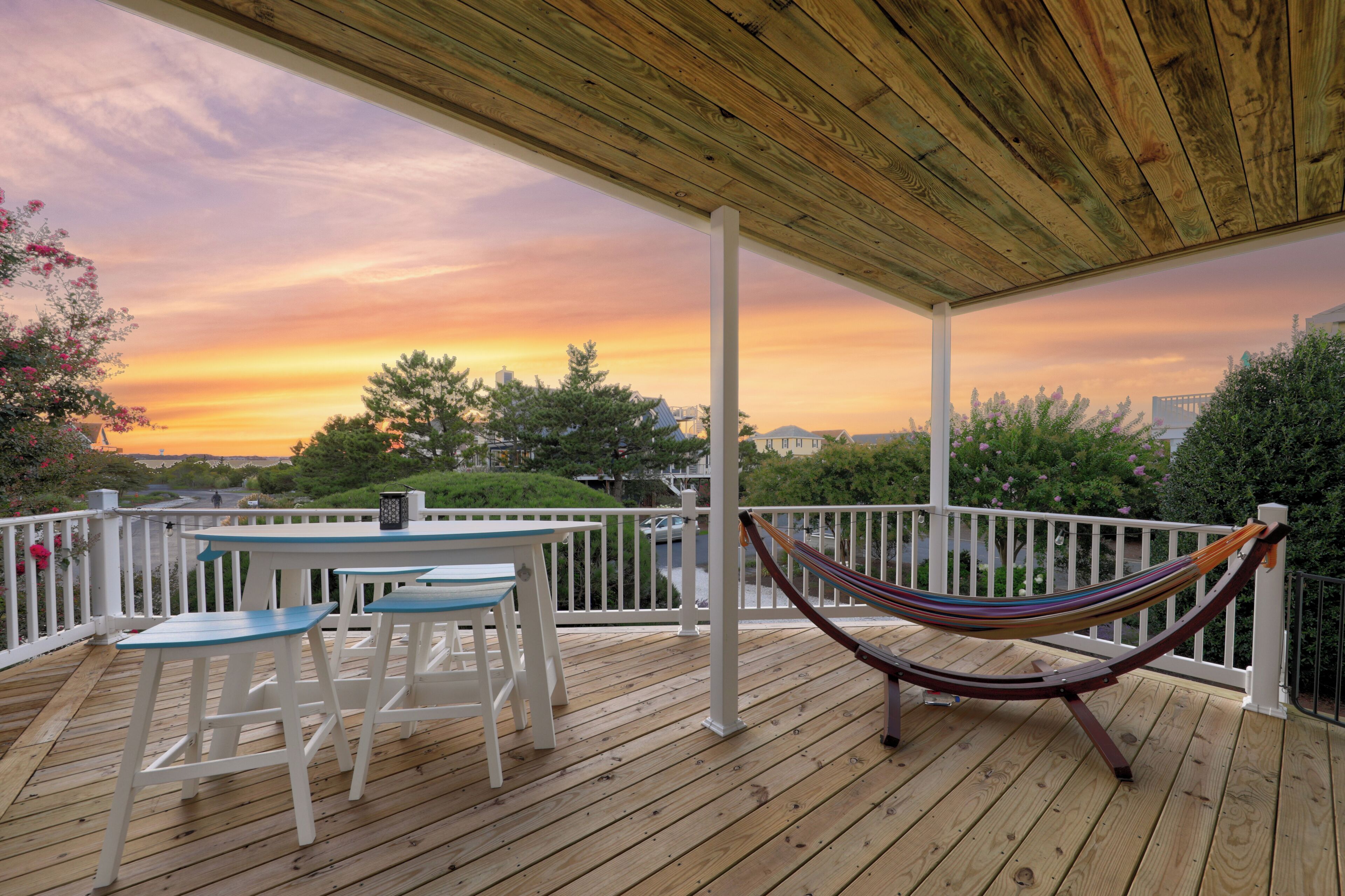 A wooden deck with a table and chairs, a hammock, and a view of the sunset.