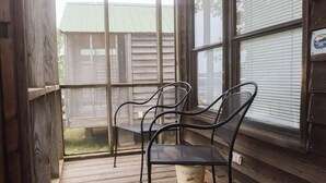 Un porche cubierto con una mesa metálica y dos sillas, suelo de madera y una ventana con persianas.