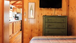 Una habitación de madera con cama, un aparador verde, televisor montado y una cocina visible a través de una puerta abierta.