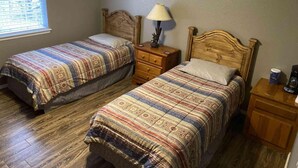 A room with two single beds, a nightstand with a lamp, a wooden dresser, and a window with blinds.