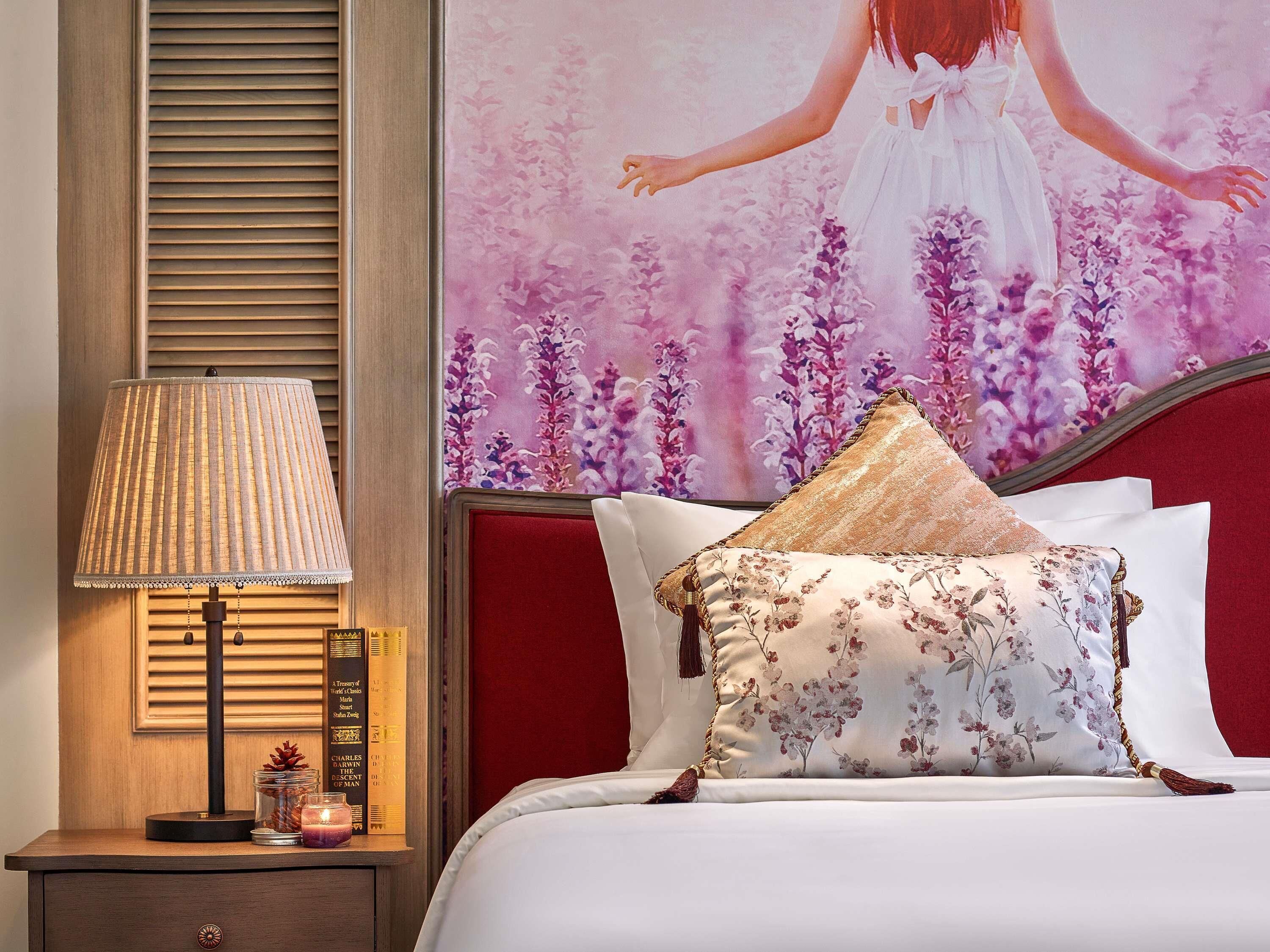 A neatly made bed with floral pillows, a bedside lamp, and a framed picture of a woman in a white dress.