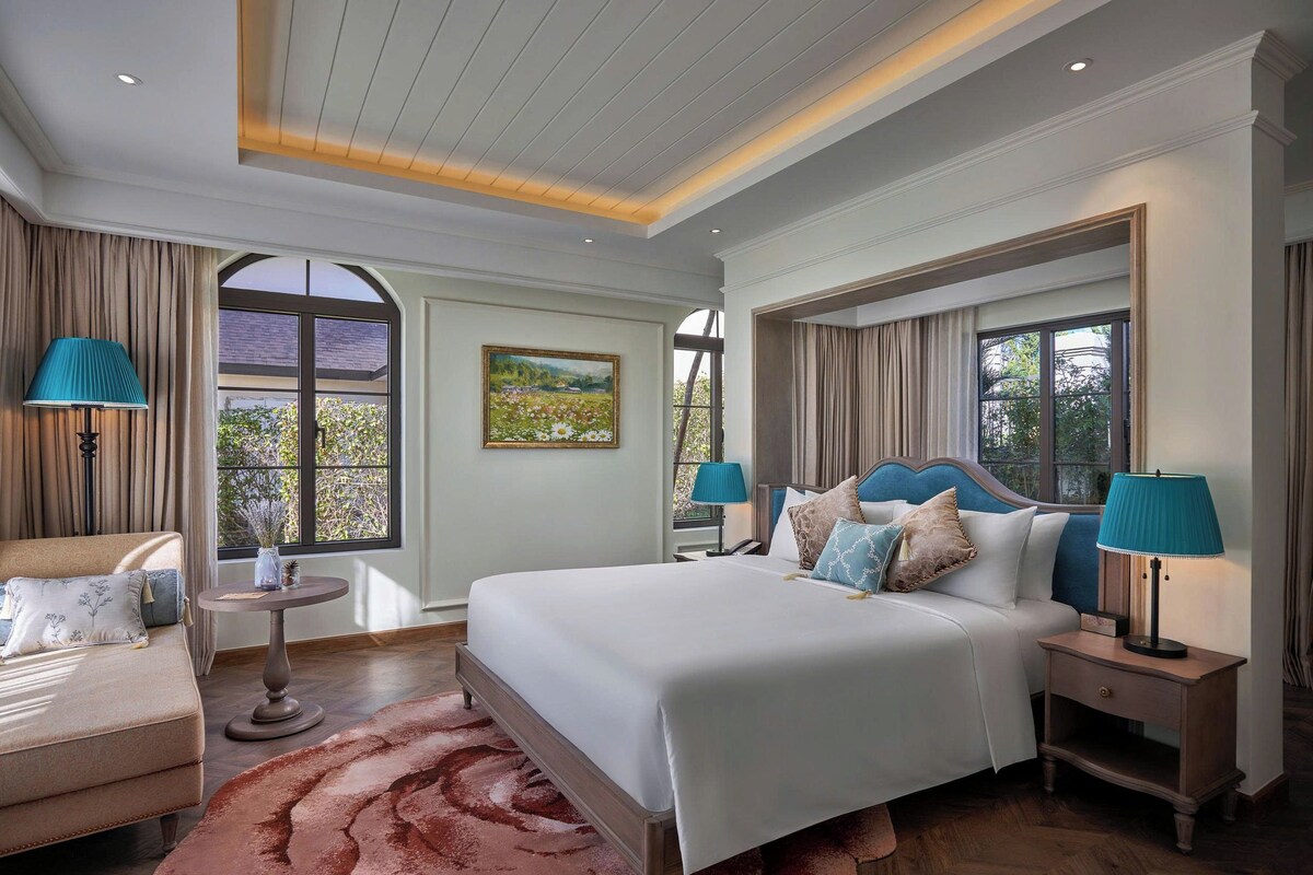 Une chambre d’hôtel avec un grand lit, deux tables de chevet, une chaise, une table ronde et une vue sur la verdure.