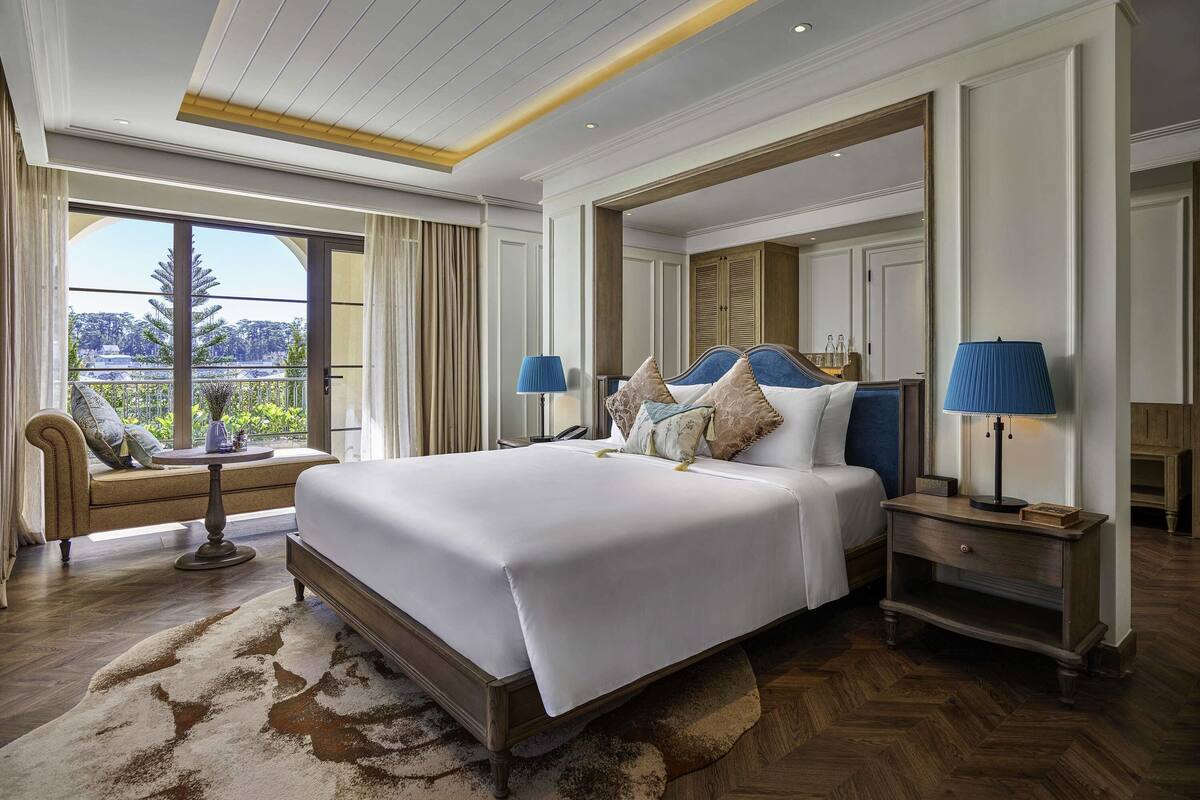 Un dormitorio espacioso con una cama grande, dos mesitas de noche, un pequeño sillón y vistas al exterior con vegetación.