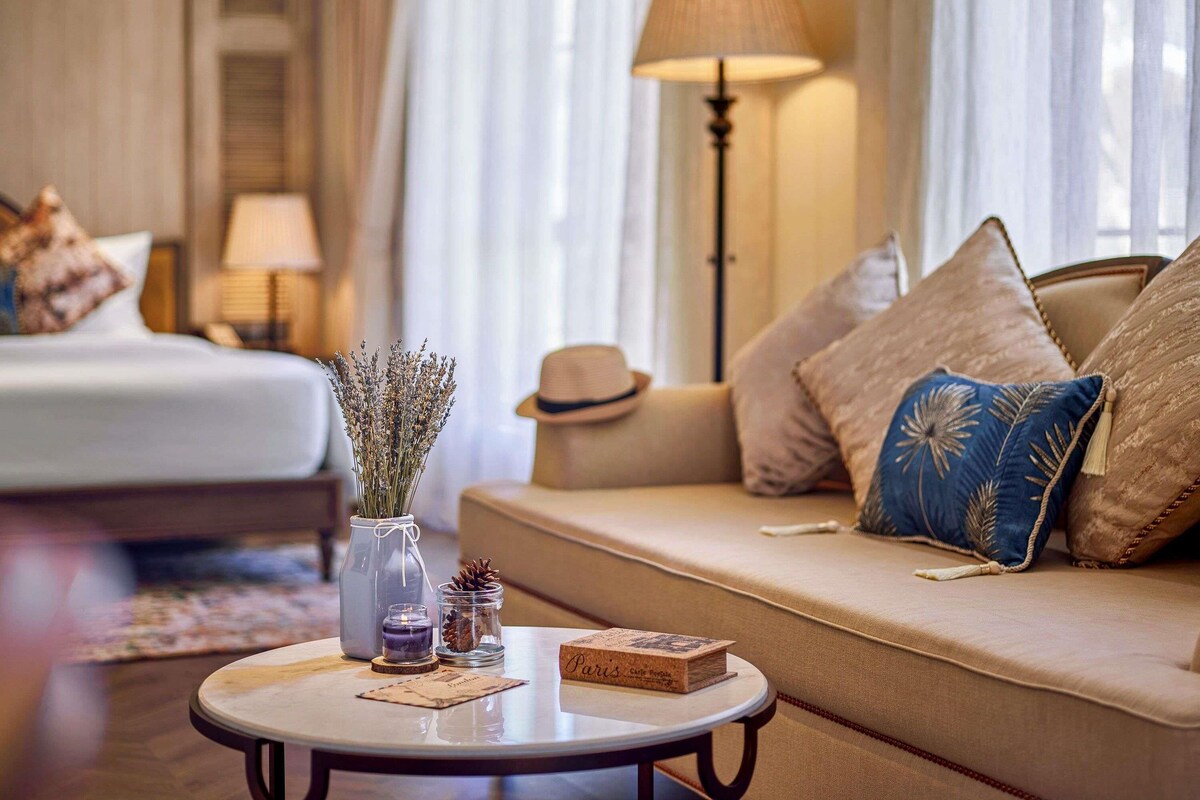 A hotel room with a bed, a sofa, a lamp, a vase with dried flowers, a candle, and a book on a side table.