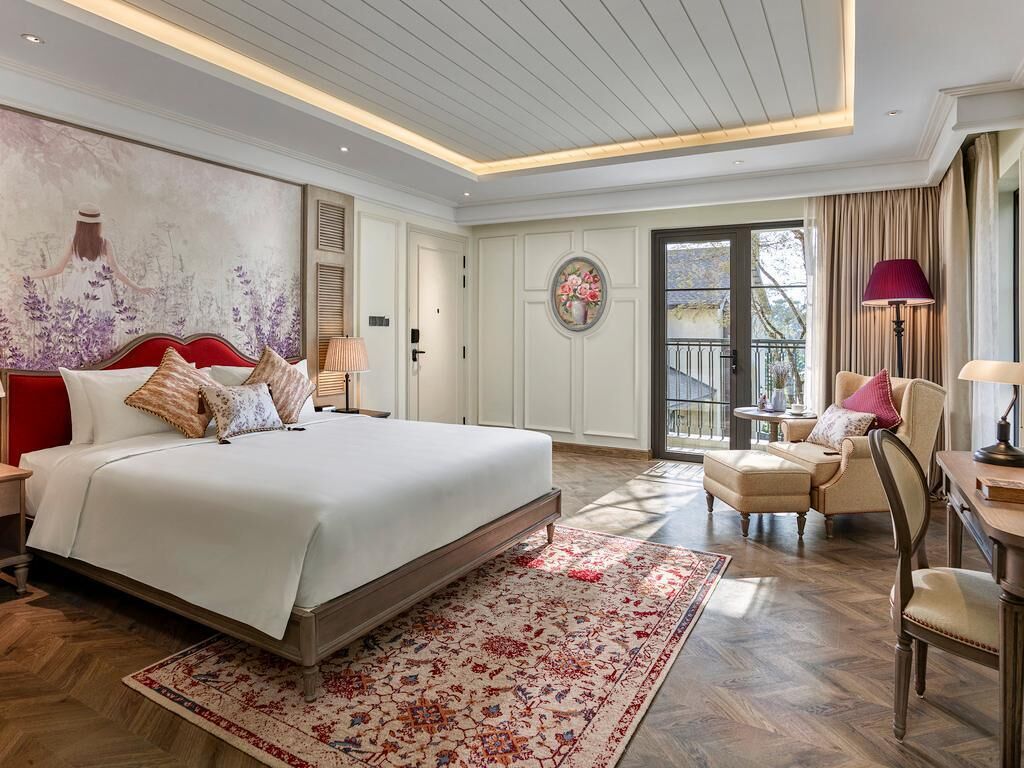 A bedroom with a large bed, a small table, a chair, a sofa, and a balcony with curtains.