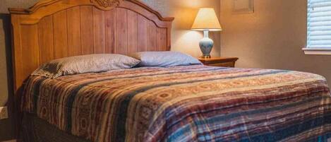A bedroom with a wooden headboard, a patterned bedspread, a bedside lamp, and a window with blinds.