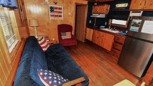 Interior acogedor de una cabaña con techo de madera, un sofá con almohadas de la bandera estadounidense, un sillón rojo, una zona de cocina con armarios de madera y un refrigerador de acero inoxidable.
