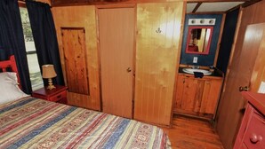 Un dormitorio de cabaña de madera con cama, mesita de noche, cómoda, espejo y un baño pequeño con lavamanos y armario.