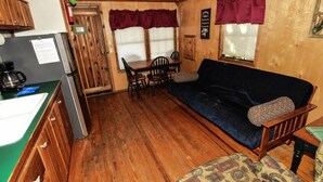 A compact kitchen with a refrigerator, sink, and wooden cabinets. A dark blue sofa and patterned ottoman are in the living area, with a wooden table and chairs. - Lake Texoma Cedar Cabin - Striper King (Mead)