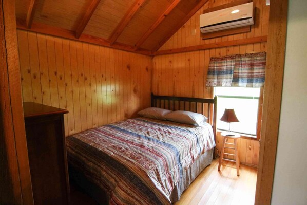 A wooden-paneled room with a bed, a window with curtains, a ladder, and an air conditioning unit.