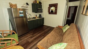 A compact kitchen with a refrigerator, microwave, and dark cabinets. A brown sofa with green cushions is in the living area, and a wicker chair is visible. - Lake Texoma Lakefront Cabin - Texoma Breeze (Mead)