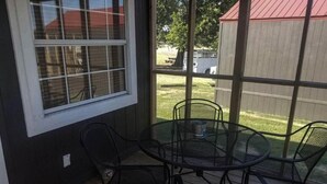 A covered patio with a round table and four chairs, a window, and a view of a backyard with a red-roofed shed. - Lake Texoma Lakefront Cabin - Texoma Breeze (Mead)
