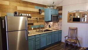 Eine kleine Küche mit einem Edelstahl-Kühlschrank, einer Mikrowelle und einem Spülbecken. Blaue Schränke säumen die Arbeitsplatte, und es steht ein Holzhocker mit gemustertem Sitzkissen bereit.