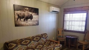 Ein Zimmer mit einer gemusterten Couch, einem kleinen Holztisch, zwei Stühlen, einem Fenster mit Jalousien und einer Wandklimaanlage.