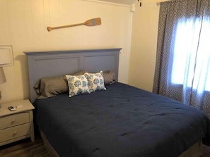 Un lit bien fait, recouvert d’une couvre-lit bleu foncé et agrémenté de deux coussins décoratifs, une décoration murale en forme de pagaie en bois, et une table de chevet avec une lampe et un téléphone.