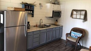 A compact kitchen with a stainless steel refrigerator, a microwave, and a sink. There is a wicker chair with a cushion, and a decorative window with a curtain.