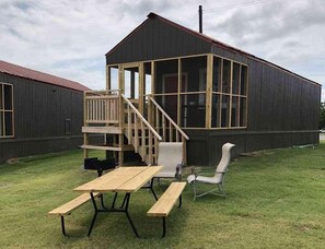 En træterrasse med et picnicbord og stole, en lille trappe samt en bygning med store vinduer og mørk facade.