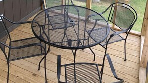 A covered patio with a round metal table and four chairs, wooden deck, and a view of a backyard with a wooden deck and chairs.