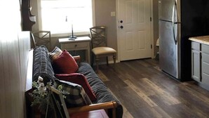 A cozy living room with a sofa, a small table, and a ceiling fan. There is a door leading to a kitchen with a refrigerator.