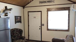 A cozy cabin interior with a wooden ceiling, a fishing-themed sign, a refrigerator, a patterned armchair, a couch, and a door.