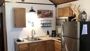 A compact kitchen with wooden cabinets, a stainless steel refrigerator, a ceiling fan, and a mounted deer head.