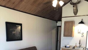 A cozy living room with a wooden ceiling, a mounted deer head, a framed picture, and a kitchenette with wooden cabinets.