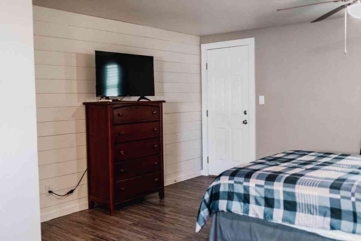 Une chambre avec une commode en bois, un téléviseur posé dessus, une couvre-lit à carreaux et une porte blanche.