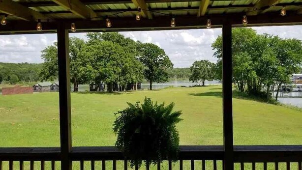 A view from a covered area with a railing, overlooking a grassy field, trees, and a body of water. - Lake Texoma Lakeside Cottage (Mead)