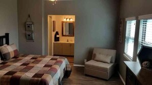 A bedroom with a bed, a chair, a nightstand, a dresser, and a bathroom with a sink and mirror. - Lake Texoma Lakeside Cottage (Mead)
