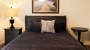Una cama con cabecera oscura, un cojín estampado, una lámpara en una mesita de noche y un cuadro enmarcado sobre la cama.