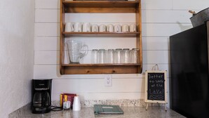 Una cocina con un estante de madera que muestra tazas blancas y una jarra de vidrio, una cafetera y una pizarra con un mensaje.