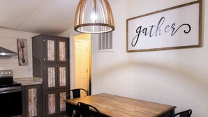 Una cocina con una mesa de comedor, sillas, una lámpara colgante y un cartel que dice 'gather' en la pared.