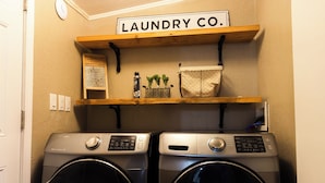Un área de lavandería con dos lavadoras de carga frontal, un estante superior con un letrero que dice 'LAUNDRY CO.' y una planta pequeña en el estante.