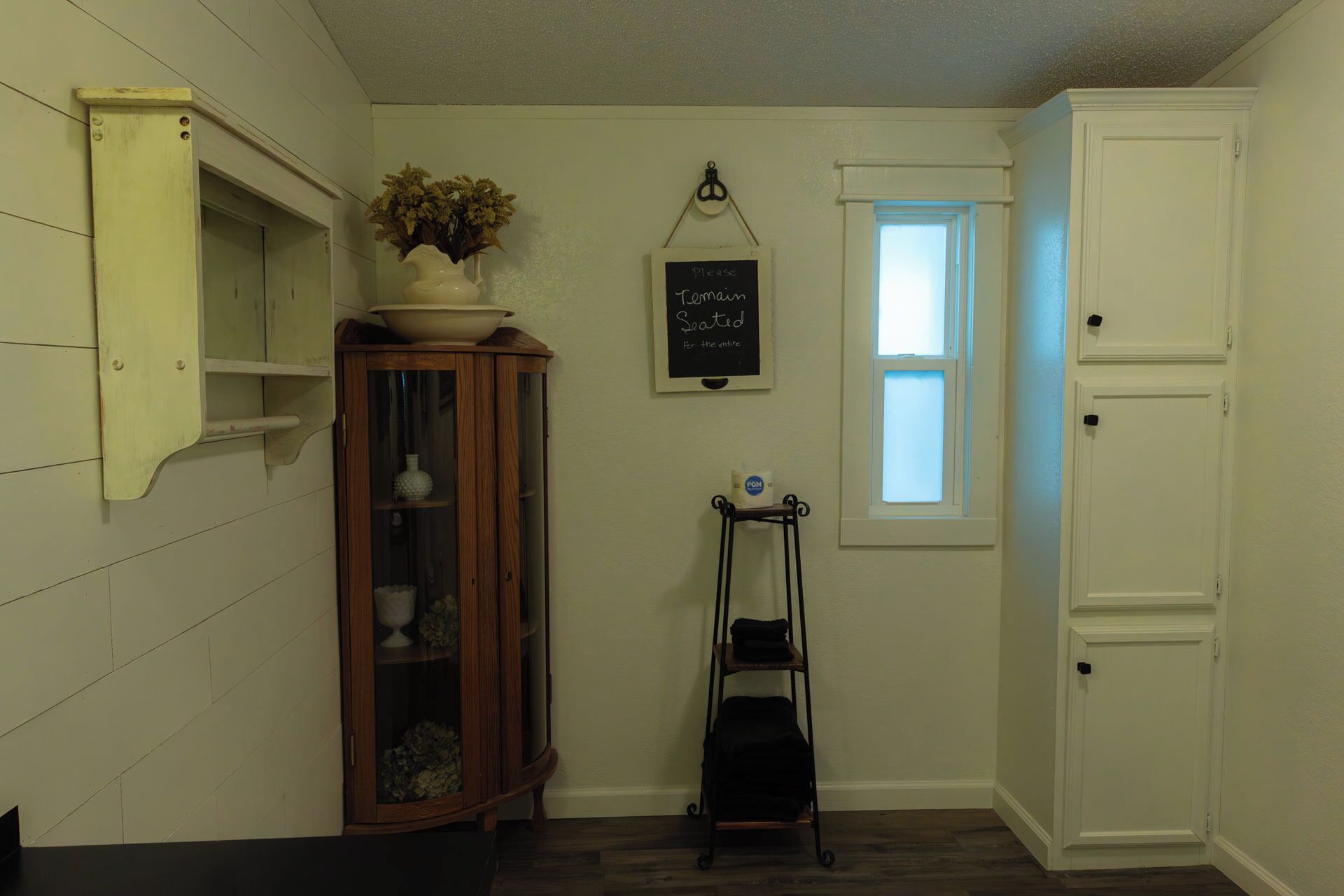 A room with a wooden cabinet, a small window, a chalkboard with a message, and a black luggage trolley.