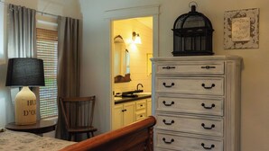 A bedroom with a bed, a nightstand, a lamp, a chair, a dresser, a birdcage, and a kitchen visible through an open door. - Lake Texoma Vacation Home The Farm House (Mead)