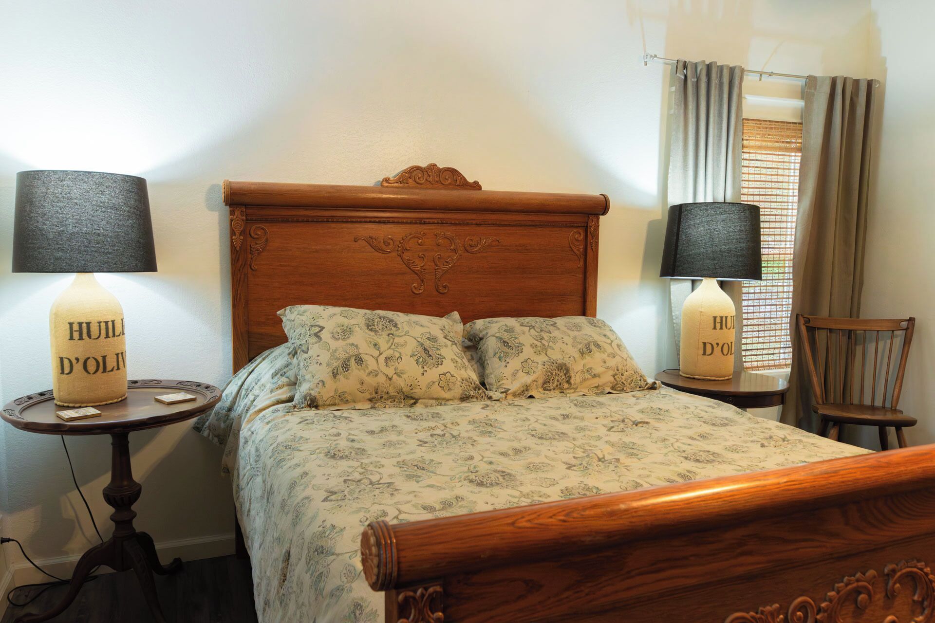 Un dormitorio con cabecera de madera, ropa de cama estampada con flores, dos lámparas de noche, una mesita y una silla.