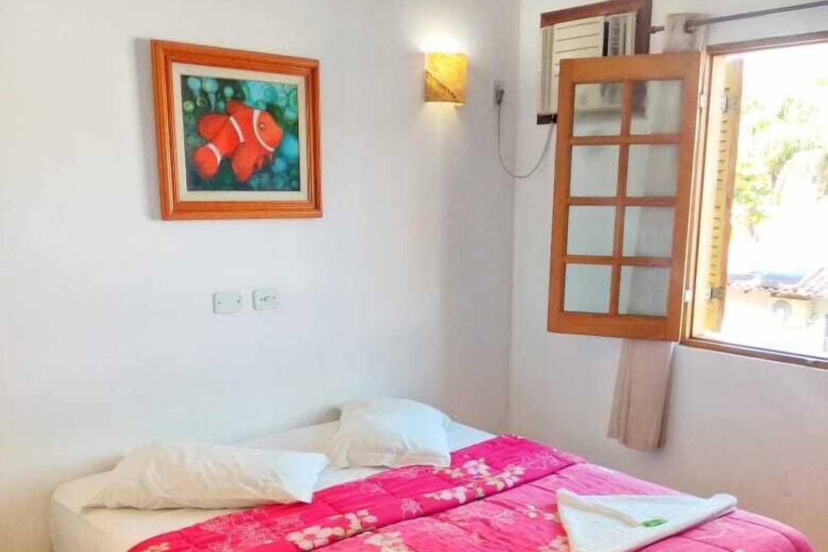 A bedroom with a bed, a painting, a wall socket, and a window with a wooden blind.