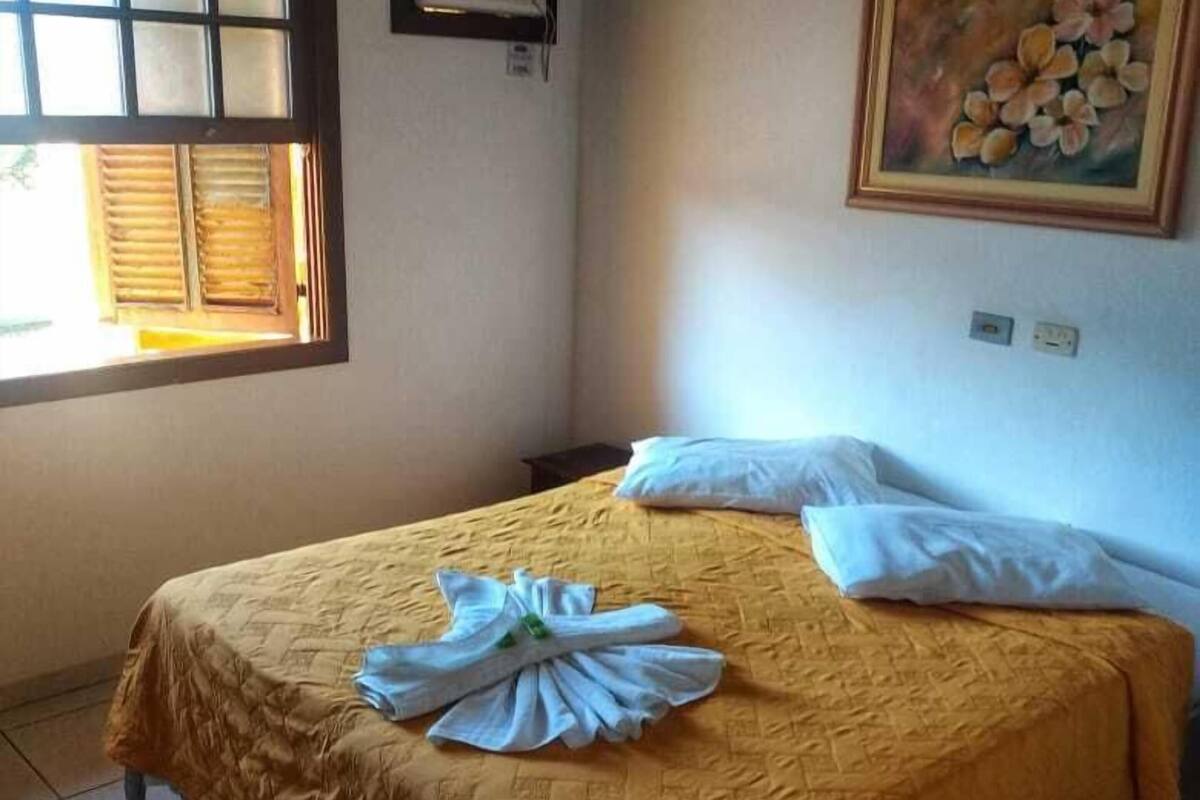 A single bed with a yellow bedspread, a window with blinds, an air conditioning unit, and a painting on the wall.