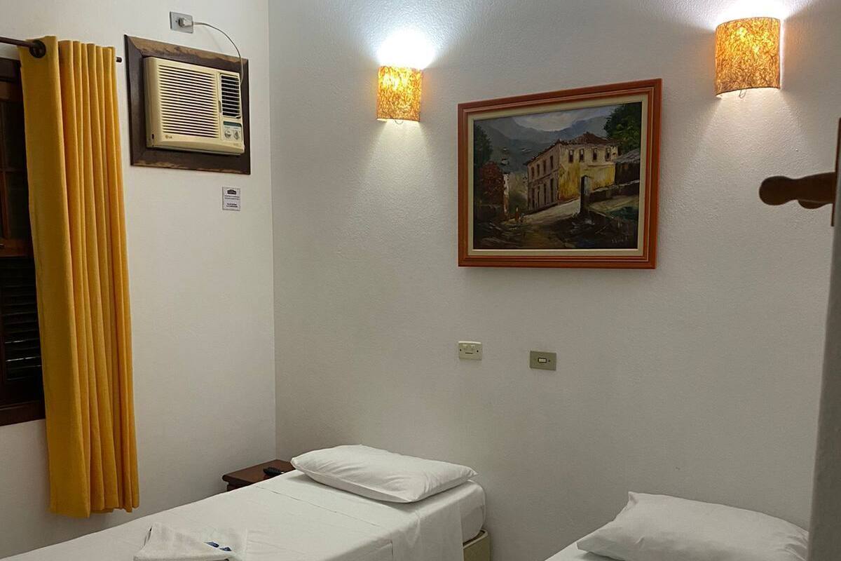 A hotel room with two single beds, a painting on the wall, a heater, and a ceiling fan.