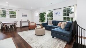 A spacious living room with a dark blue sofa, a round ottoman, and a dining area with a wooden table and chairs. - Crestwood Contemporary - Minutes to Downtown! (Birmingham)