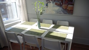 A dining table with white chairs and green placemats, a vase with green plants, and a painting on the wall.