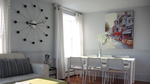 A modern living room with a sofa, a dining table with chairs, a clock on the wall, and a painting.