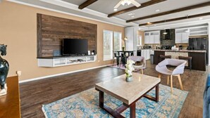A spacious living room with a large TV, wooden beams, a ceiling fan, and a kitchen area visible in the background. - 8 acres Ranch - 2 Homes - Sleeps 16 - Dripping (Dripping Springs)