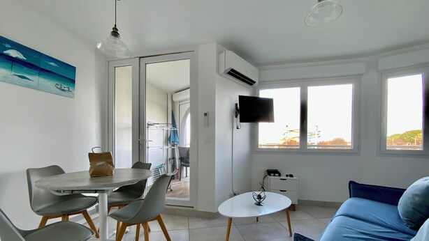 Een moderne woonkamer met een blauwe bank, een eettafel met stoelen, een aan de muur bevestigde televisie en grote ramen.