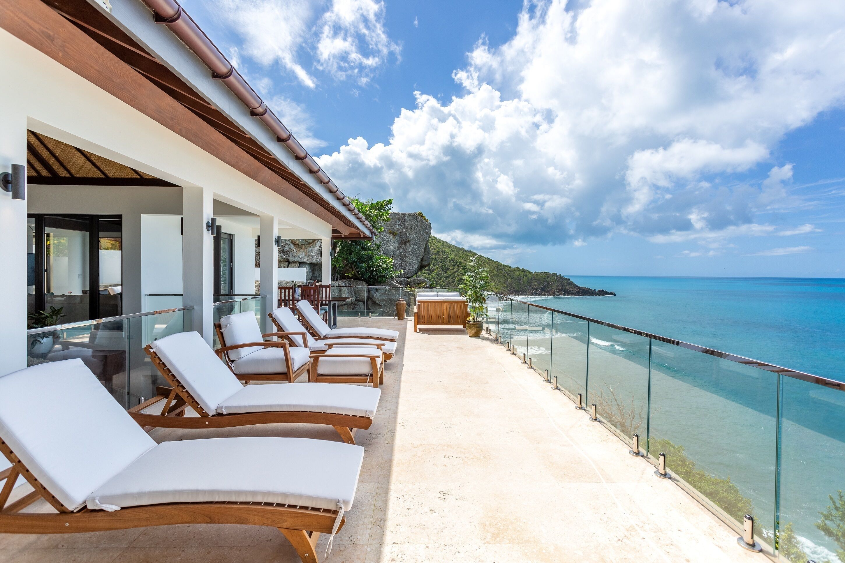 A balcony with sun loungers overlooking a clear blue sea, a rocky outcrop, and a white building with a wooden roof.