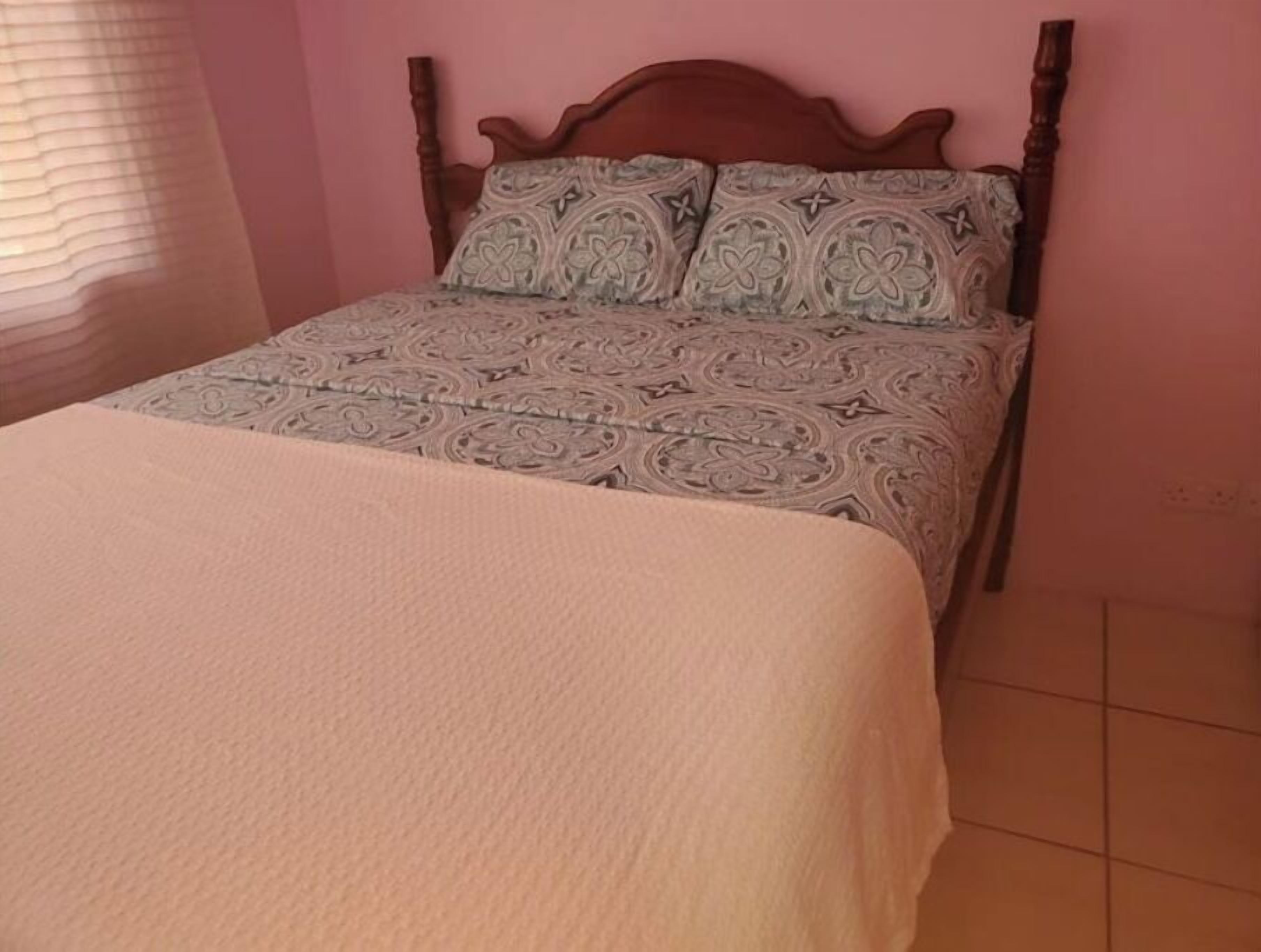 Una cama con colcha y almohadas estampadas, cabecera de madera y ventana con persianas.