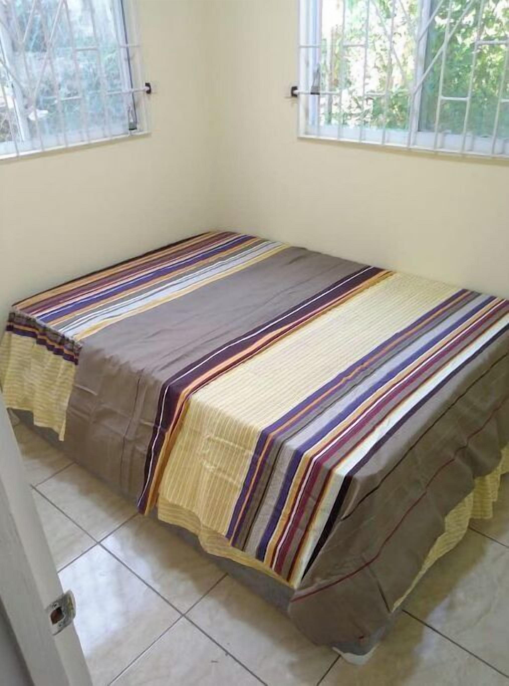 Un lit simple avec une couvre-lit à rayures, dans une pièce dotée de deux fenêtres et d’un sol carrelé.