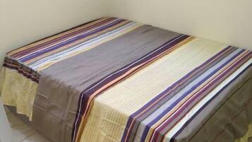 Un lit simple avec une couvre-lit à rayures, dans une pièce dotée de deux fenêtres et d’un sol carrelé.