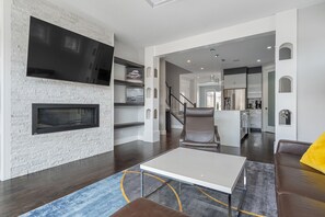 A modern living room with a fireplace, a flat-screen TV, a dining area, and a kitchen in the background.